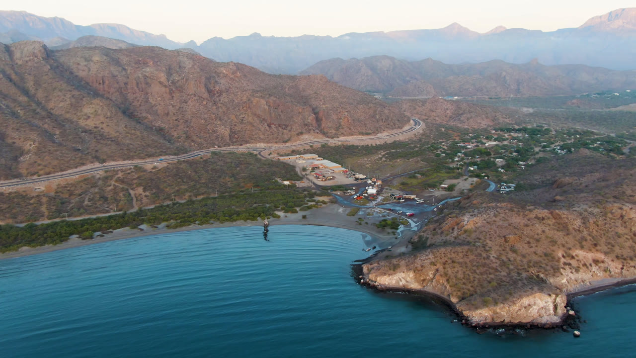 Aerial frame over Danzante Bay in Loreto, Baja California Sur, with turquoise water in front, a curved beach, a rocky headland, a road, scattered buildings, and no visible people.