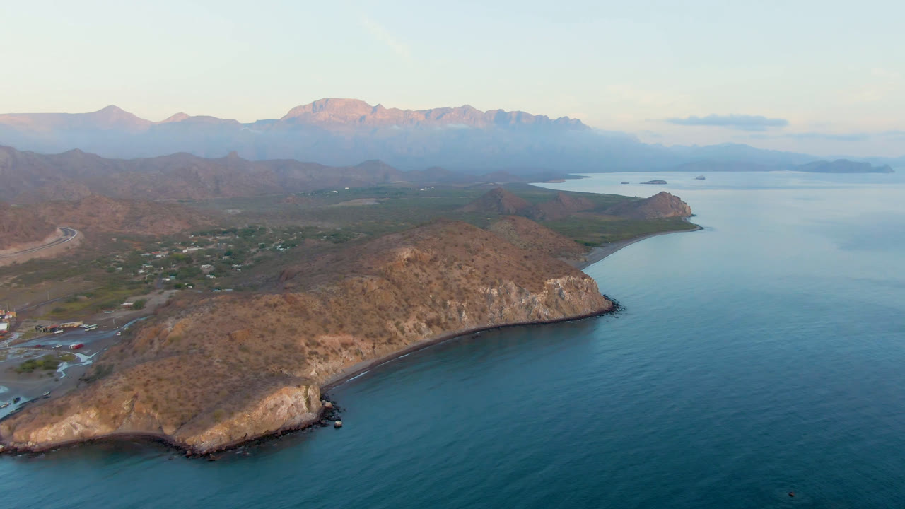 Aerial frame over Danzante Bay Loreto showing blue water below a rocky headland, a small shoreline at left, hazy mountains beyond, and no visible people.
