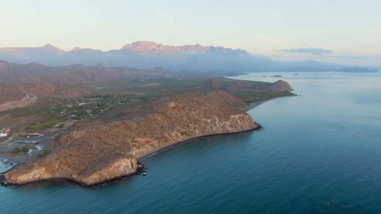 Wide aerial view of Danzante Bay Loreto with the rocky point centered, calm blue water on the right, small shore activity at left, and no visible people.