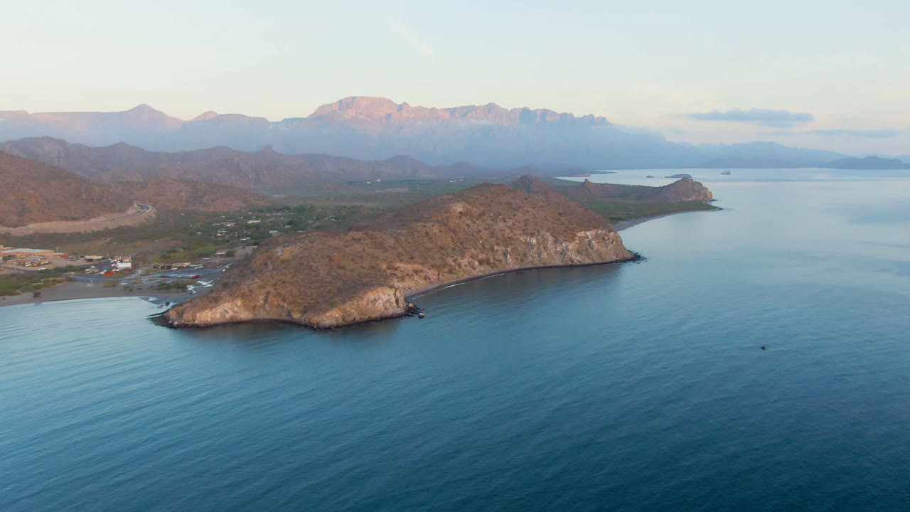 Wide aerial view of a rocky island, blue ocean, coast, and distant mountains.