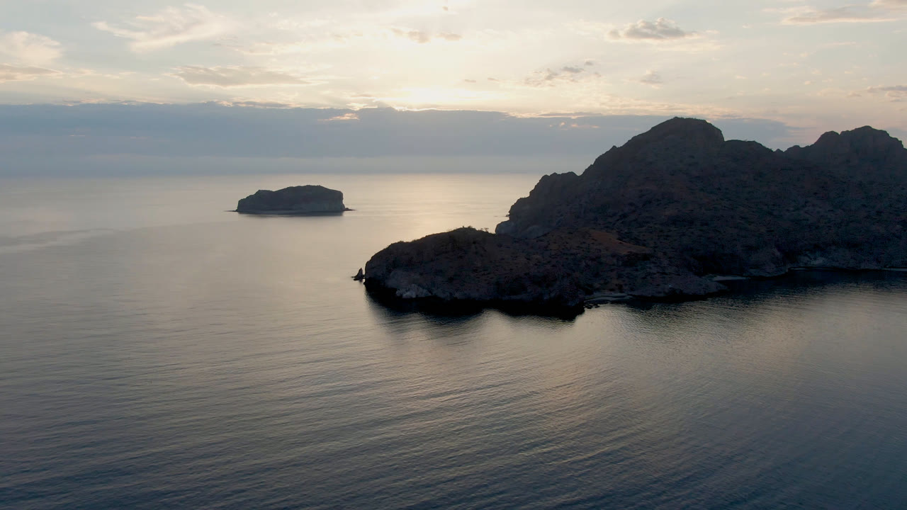 Opening aerial view of Danzante Bay Loreto with a small island left of center, dark rocky coastline on the right, calm ocean in front, and no visible people.