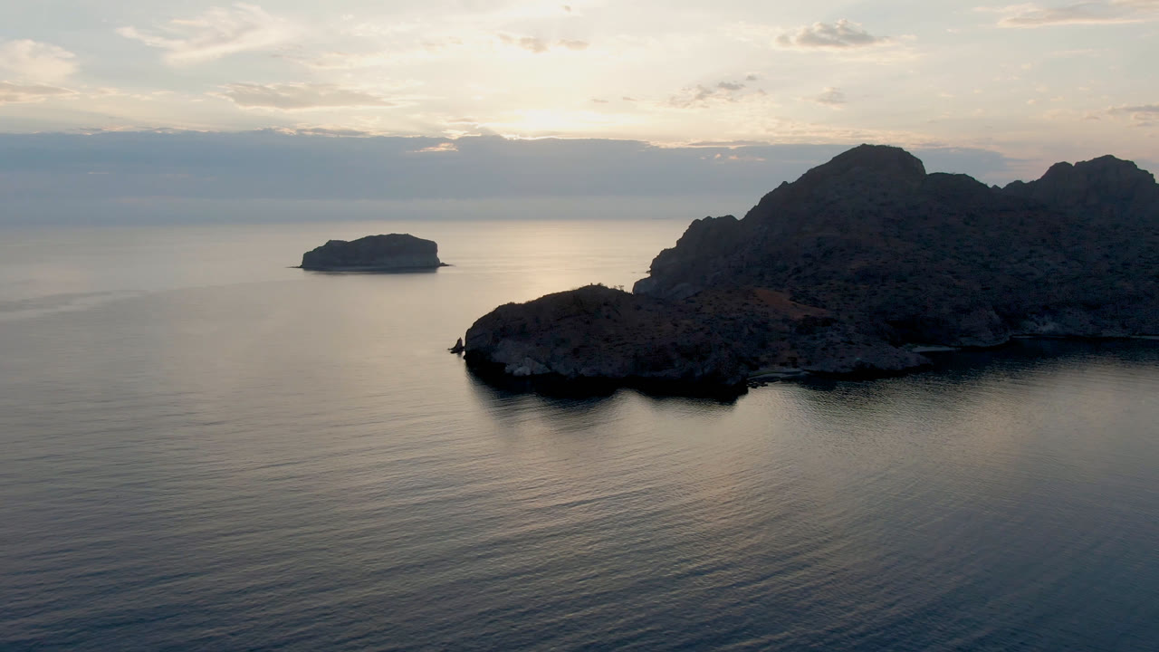Wide Danzante Bay Loreto frame where the small island sits against calm water, the rugged coast stays to the right, layered clouds soften the horizon, and no visible people appear.