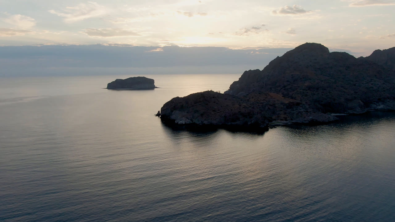 Aerial frame from Danzante Bay near Loreto showing a pale reflection path on calm water, a small left-side island, darker rocky land to the right, and no visible people.