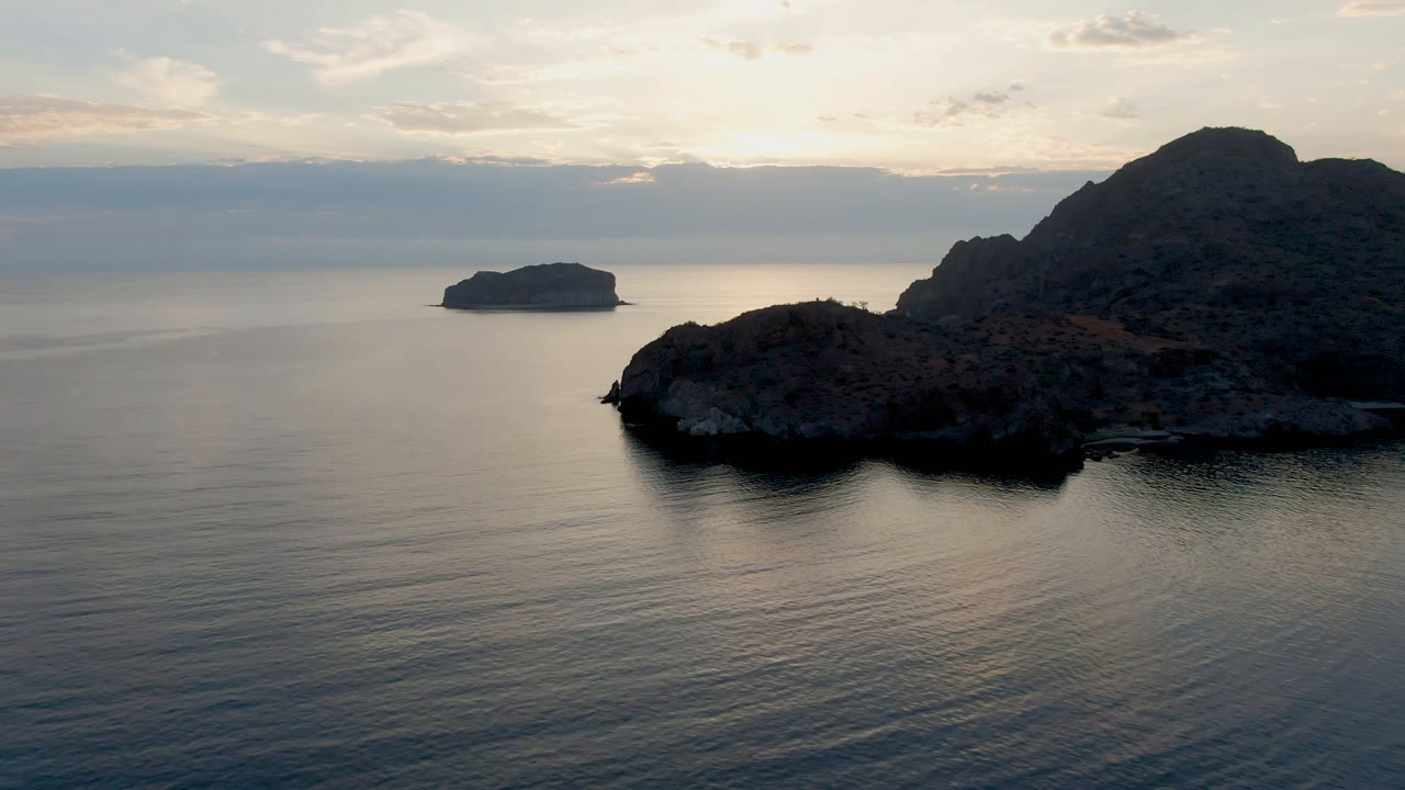 Wide aerial view of Danzante Bay in Loreto, Baja California Sur, with dark rocky shoreline, calm open water, a small distant island, layered clouds, and no visible people.