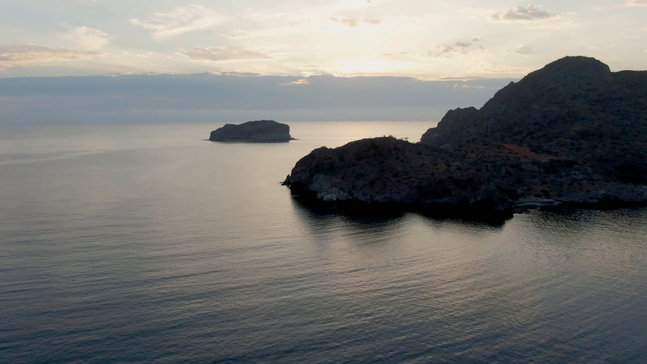 Aerial frame from Danzante Bay showing the rocky point shifted slightly across calm water, with the small island left of center, pale cloud cover, and no visible people.
