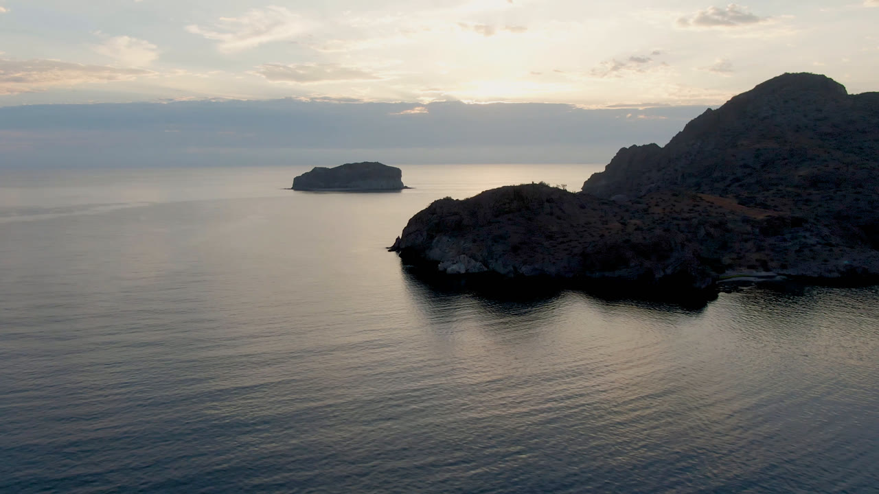 Danzante Bay aerial still with the rocky shoreline occupying the right side, smooth water in the foreground, a small island near the left horizon, soft clouds, and no visible people.