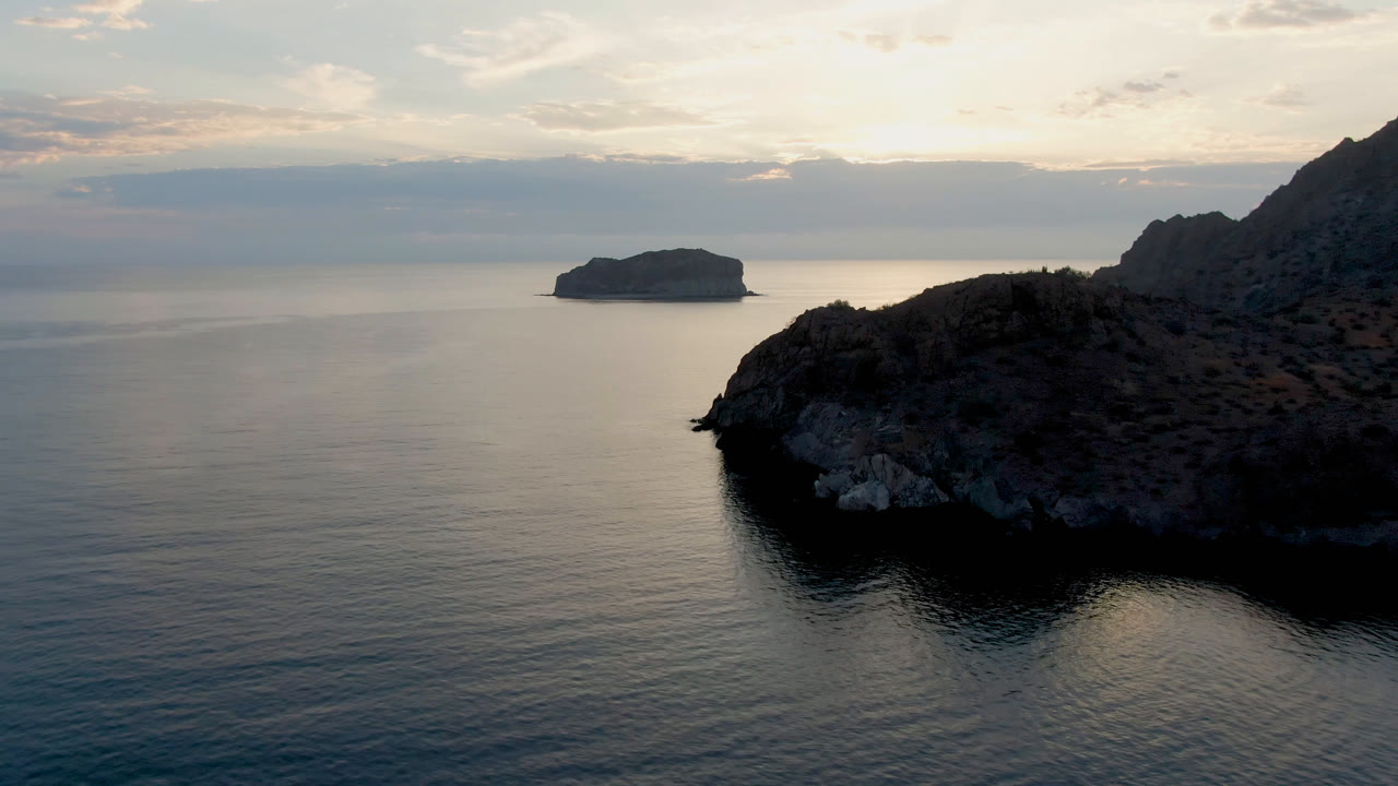 Opening view of Danzante Bay in Loreto, Baja California Sur, with a dark rocky shore at right, calm water, a distant island, soft cloud light, and no visible people.