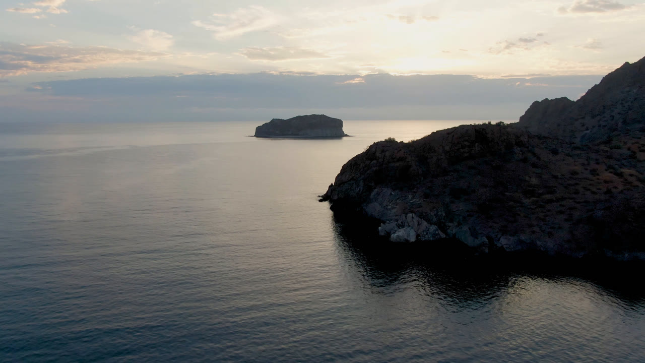 One second into the Danzante Bay clip, the rocky headland remains prominent on the right while blue-gray water, a low island, and a clouded horizon fill the left side, with no visible people.