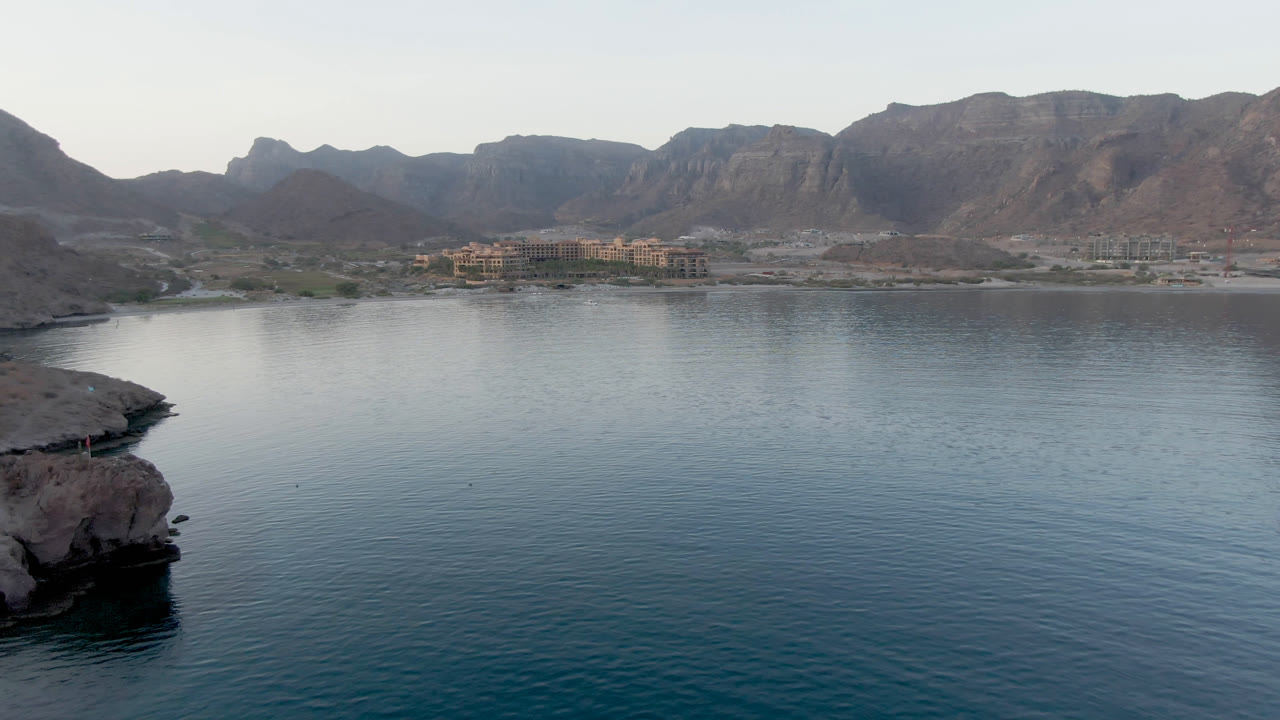 Wide view of Danzante Bay in Loreto, Baja California Sur, with calm water in the foreground, a rocky edge at left, distant shoreline buildings, mountains, and no visible people.