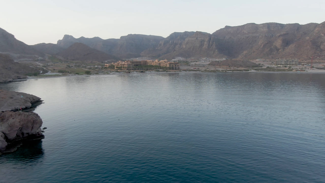 Danzante Bay water fills the lower frame as a rocky point projects near center, with shoreline buildings and a broad mountain ridge behind them, and no visible people.