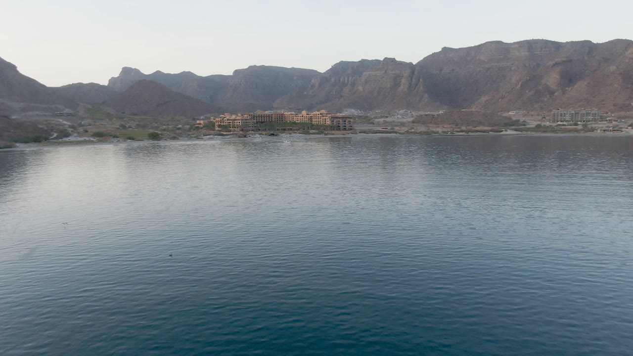One second into the Danzante Bay clip, calm bay water occupies most of the frame while the shoreline buildings and mountain ridge sit under a clear pale sky with no visible people.