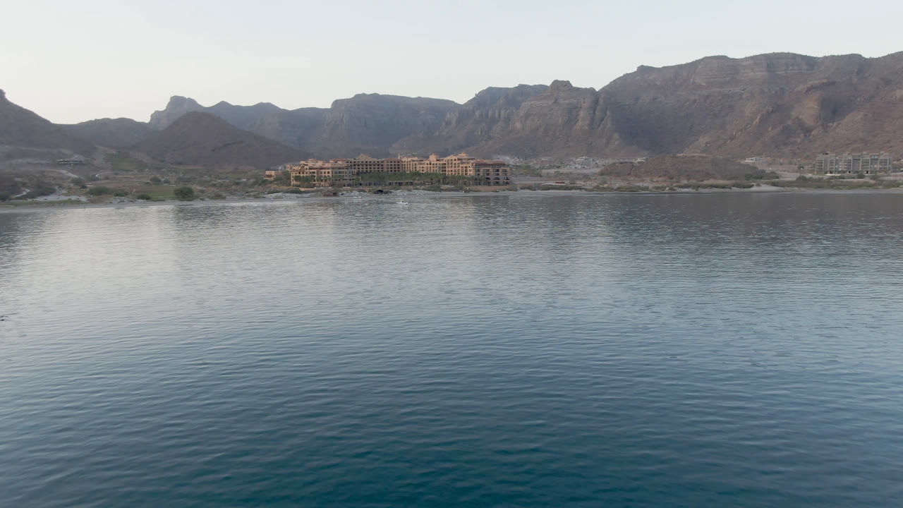 Opening frame of Danzante Bay in Loreto, Baja California Sur, with rippled water in the foreground, small shoreline buildings, brown mountains, pale sky, and no visible people.