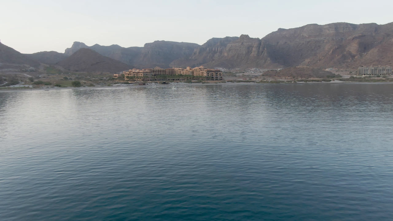 Early frame in Danzante Bay, Loreto, showing blue-green water with fine ripples, a developed shoreline, layered brown mountains, light sky, and no visible people.