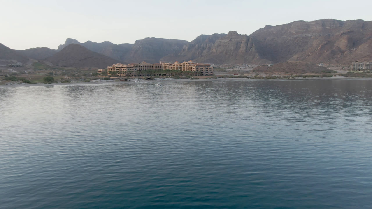 Opening frame of Danzante Bay Loreto with rippled blue water in front, low shoreline buildings, rugged mountains in the background, and no visible people.