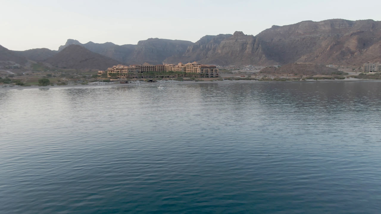 Danzante Bay Loreto view with a broad reflective water foreground, clustered shoreline buildings near center, brown mountain ridges behind, and no visible people.