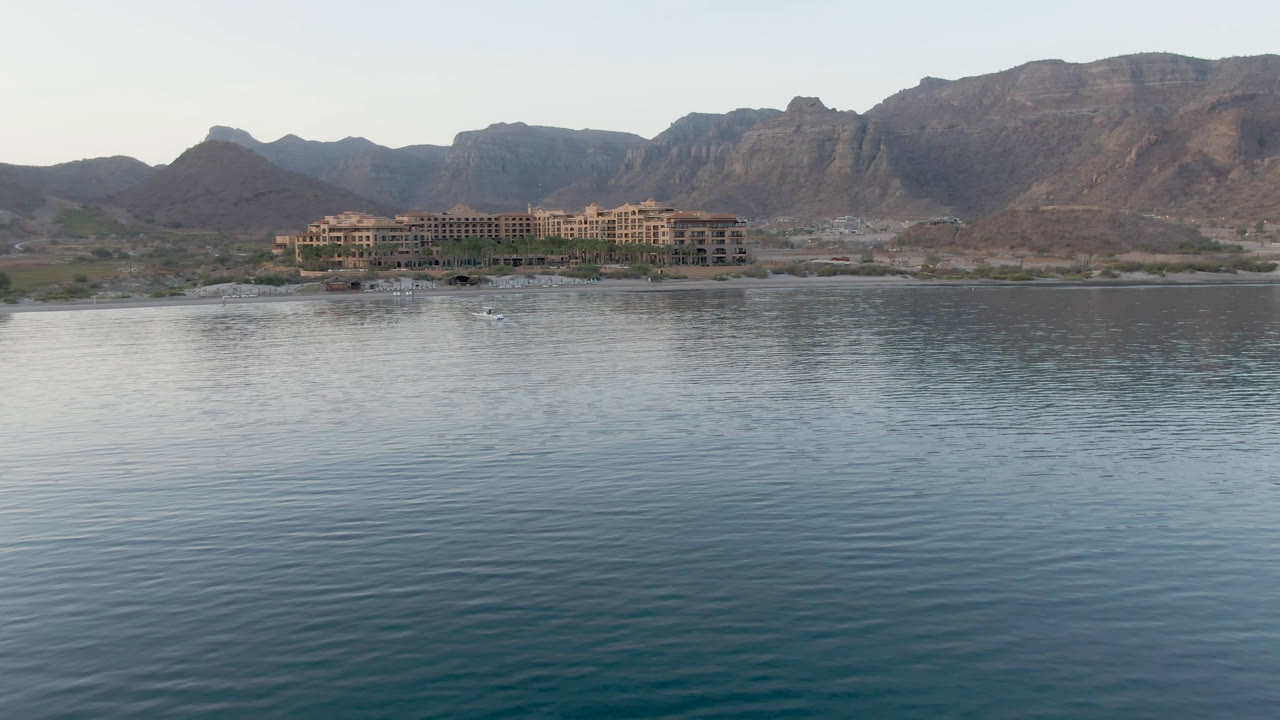 Loreto, Mexico resort shoreline seen across smooth blue-green water, with layered brown mountains across the background and no visible people.
