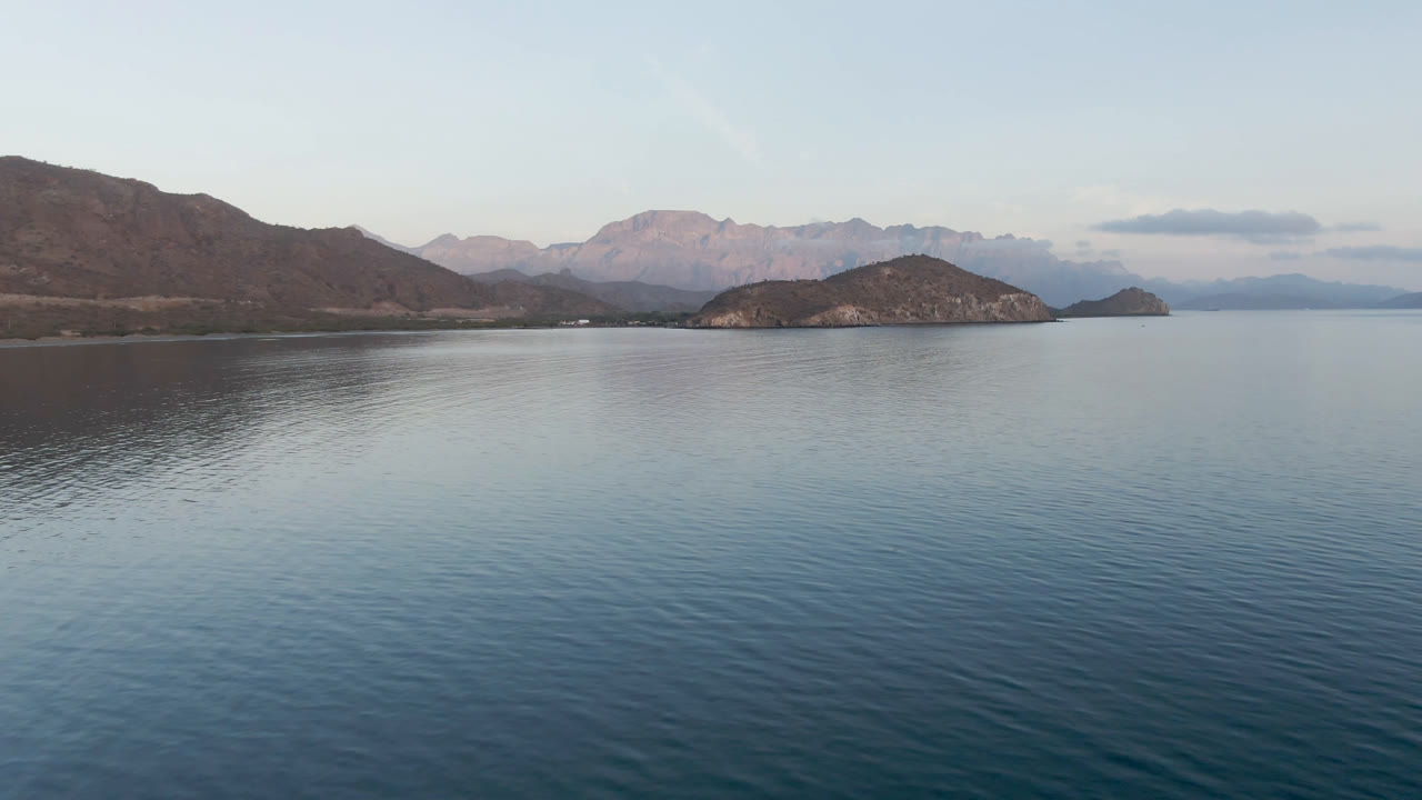 Opening frame of Danzante Bay in Loreto, Baja California Sur, with rippled blue water in front, dark shoreline at left, rocky islands near center, distant mountains, pale sky, and no visible people.