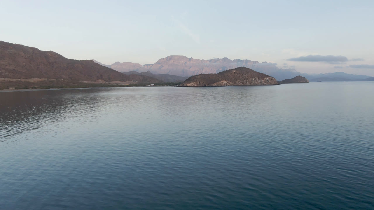 At 0:02 in Danzante Bay, the frame holds a wide elevated view over textured blue water toward the rocky island group, with Baja California Sur mountains in haze and no visible people.