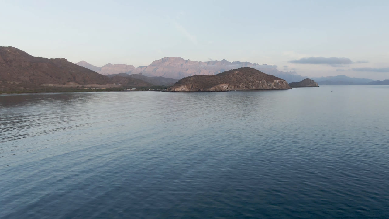 Wide aerial frame of Danzante Bay in Loreto, Baja California Sur, with calm blue water in front, rocky island shapes near the horizon, distant mountains, and no visible people.