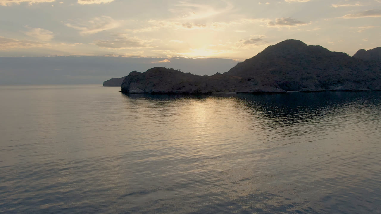 Danzante Bay water fills the foreground while dark rocky land and a pale sun reflection sit beneath layered clouds, with no visible people.