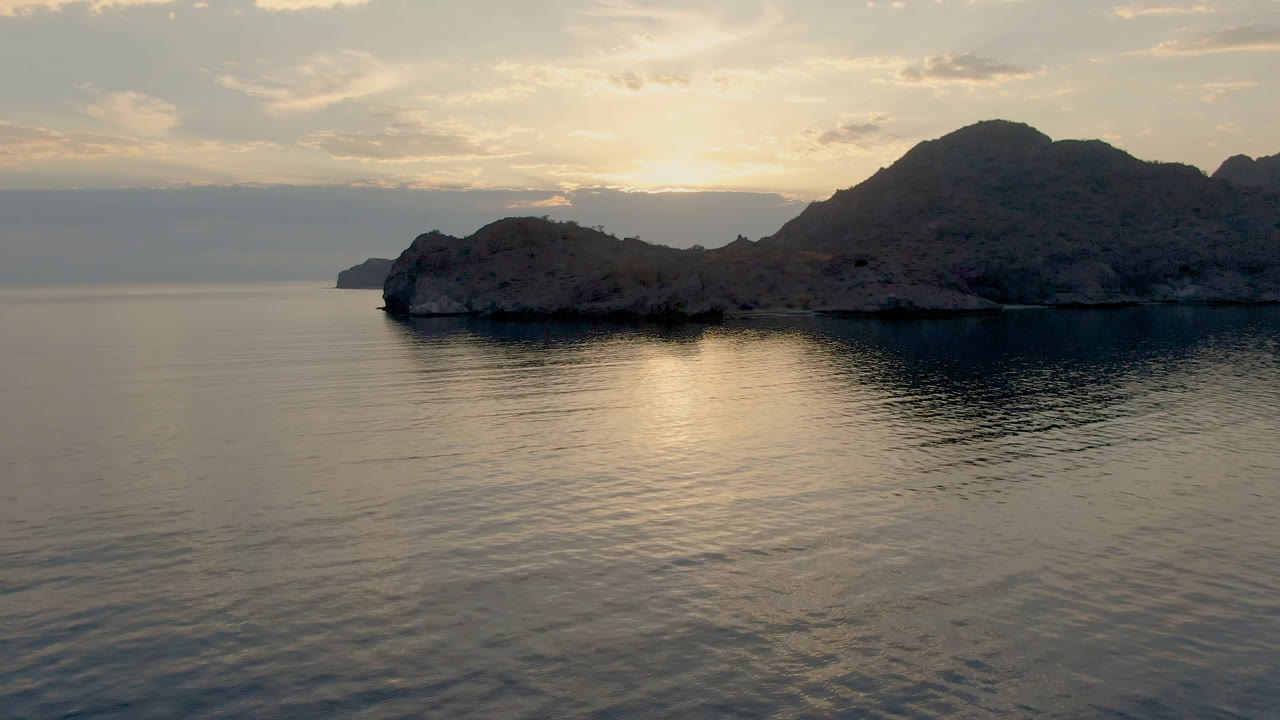 Wide view of Danzante Bay Loreto with calm rippled water in front, a rocky island and dark mountain ridge under soft clouded sunset light, and no visible people.