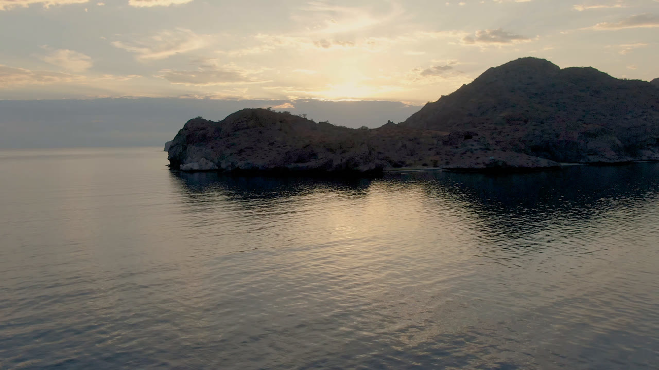 Opening frame of Danzante Bay in Loreto, Baja California Sur, with calm water reflecting pale sunset light below a rocky island and dark mountain silhouette, no visible people.