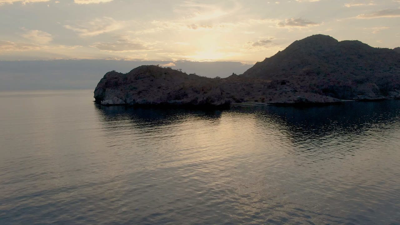 Second frame of Danzante Bay in Loreto, Baja California Sur, with the rocky island slightly left of center, the mountain ridge dark against a pale sky, and no visible people.