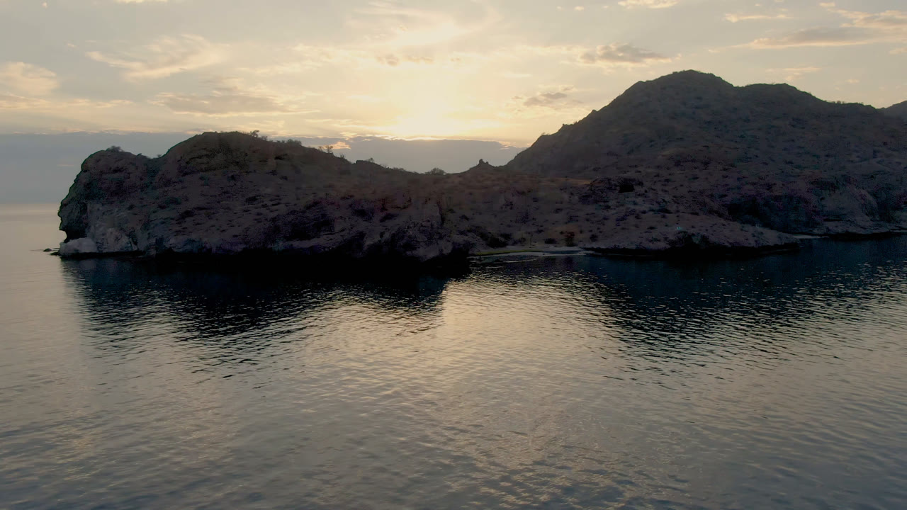 Danzante Bay coastline appears in an elevated wide frame, with rippled water reflecting the pale sky, rocky slopes in shadow, a larger mountain behind them, and no visible people.