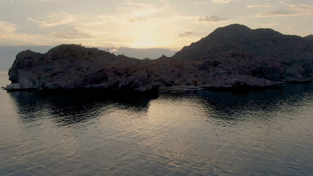 A wide view over Danzante Bay shows a dark rocky island shape against reflective water, mountain ridges in the background, a bright clouded horizon, and no visible people.