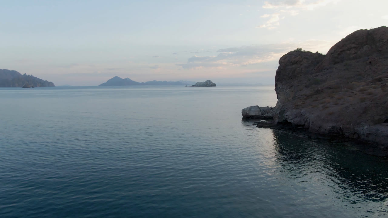 Second frame of Danzante Bay Loreto showing the rock face slightly more central, glassy water below, pale sky above, distant islands, and no visible people.