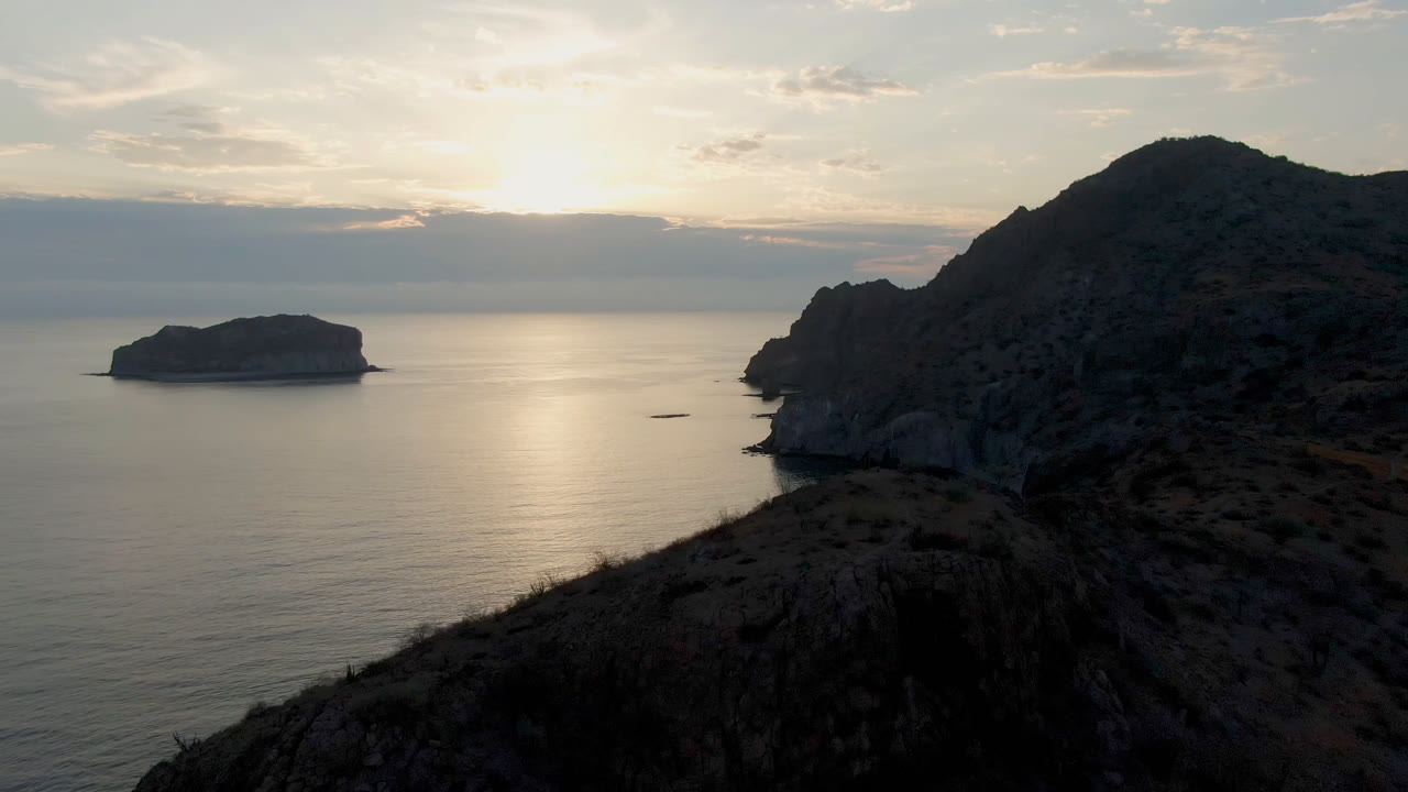Opening frame of Danzante Bay Loreto with a dark rocky headland at right, a low offshore rock at left, sunlit water, scrub foreground, and no visible people.