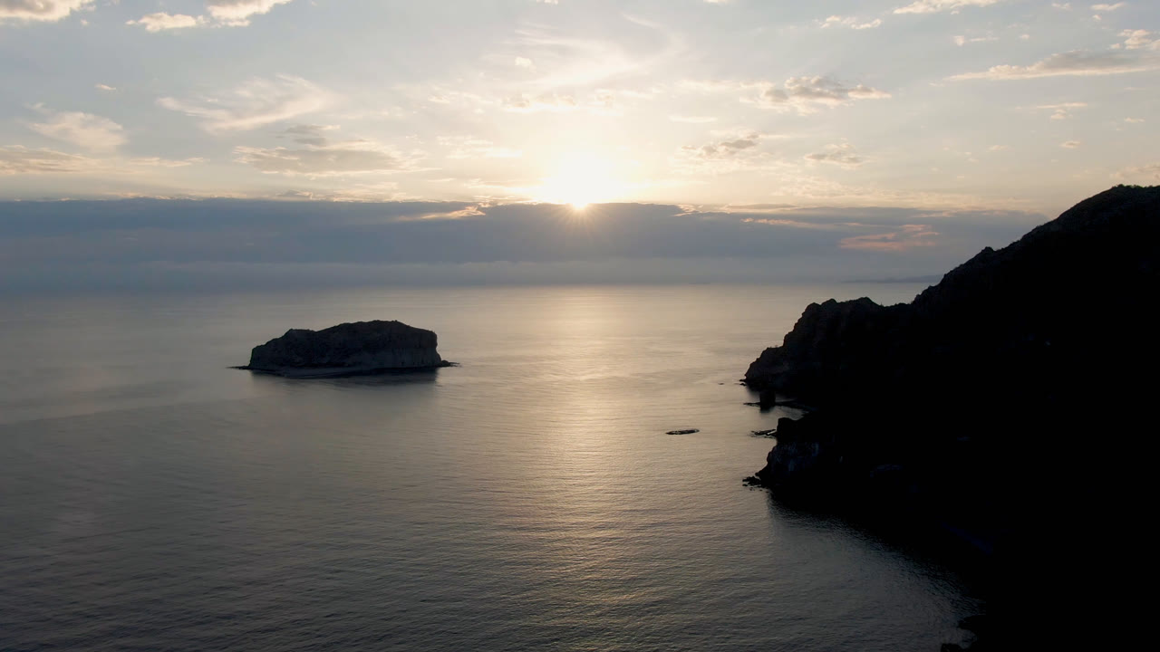 Small island in calm ocean beside a dark headland at low sun.