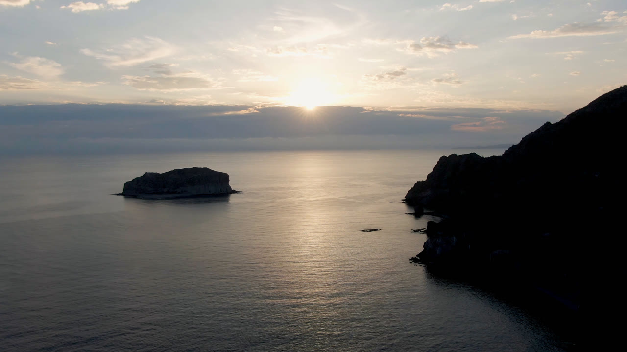 Danzante Bay Loreto ocean view at sunset with a low island rock left of center, a dark cliff on the right edge, cloud bands on the horizon, and no visible people.