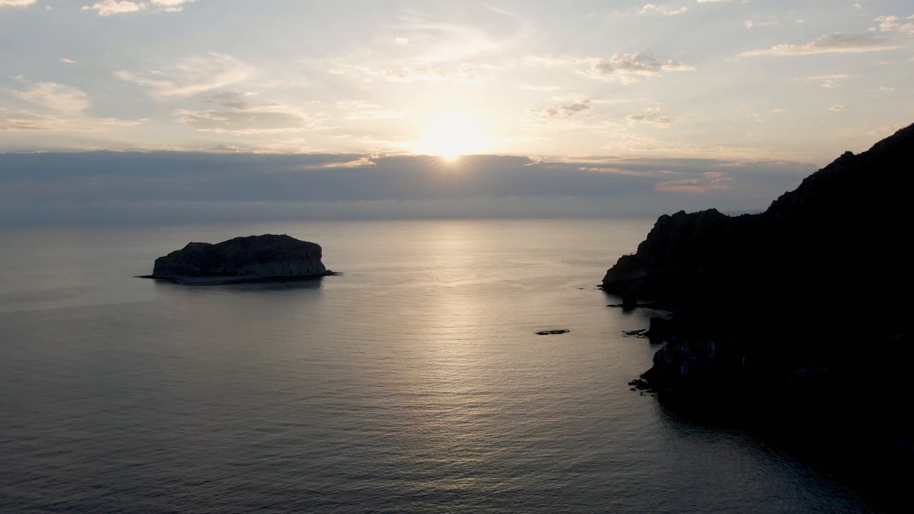 Danzante Bay, Loreto, Baja California Sur frame showing a small island left of center, low sun behind clouds, calm ocean, and a dark cliff edge at right with no visible people.