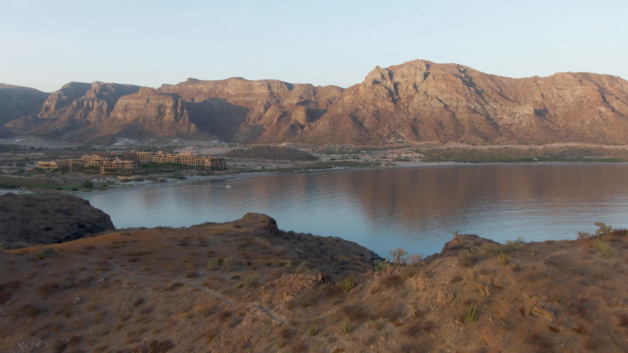 Opening frame of Danzante Bay Loreto with dry rocky foreground, calm water, distant shoreline buildings, sunlit mountains, pale sky, and no visible people.
