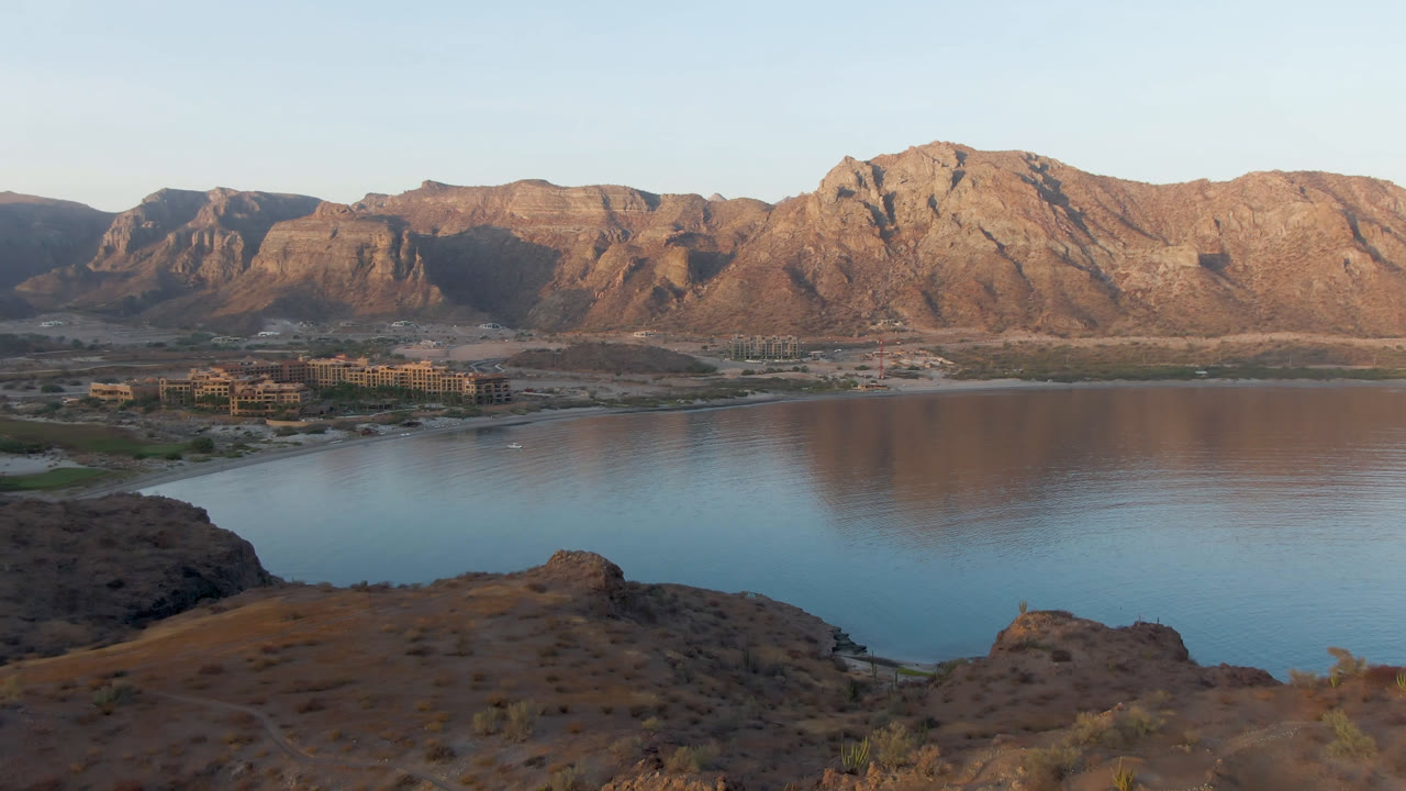 Wide view of Danzante Bay Loreto with calm blue water, rocky foreground slopes, tan mountains in warm light, and no visible people.