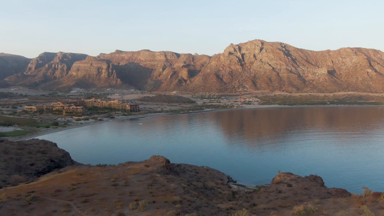 Danzante Bay Loreto seen from above with a broad band of water, small distant shoreline buildings, warm mountain ridges, and no visible people.