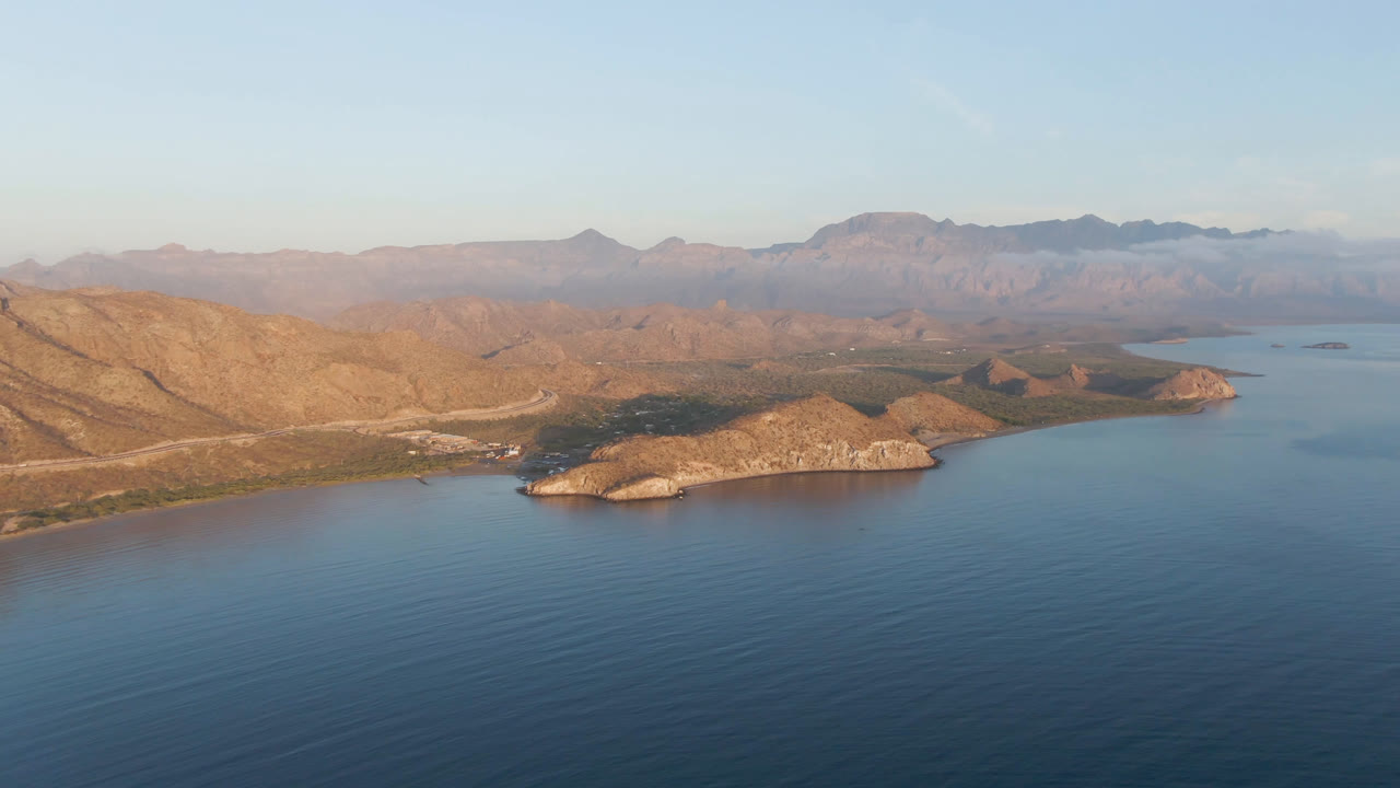 Wide aerial frame over Danzante Bay Loreto, with dark blue water in the foreground, a rocky point across the center, tan hills behind it, hazy mountains on the horizon, and no visible people.