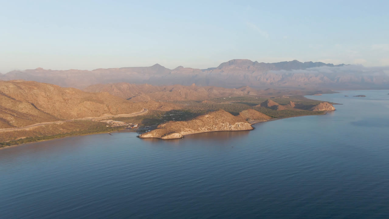 A second aerial frame of Danzante Bay Loreto shows calm blue water, a sunlit rocky headland near the center, muted desert ridges in back, pale sky, and no visible people.