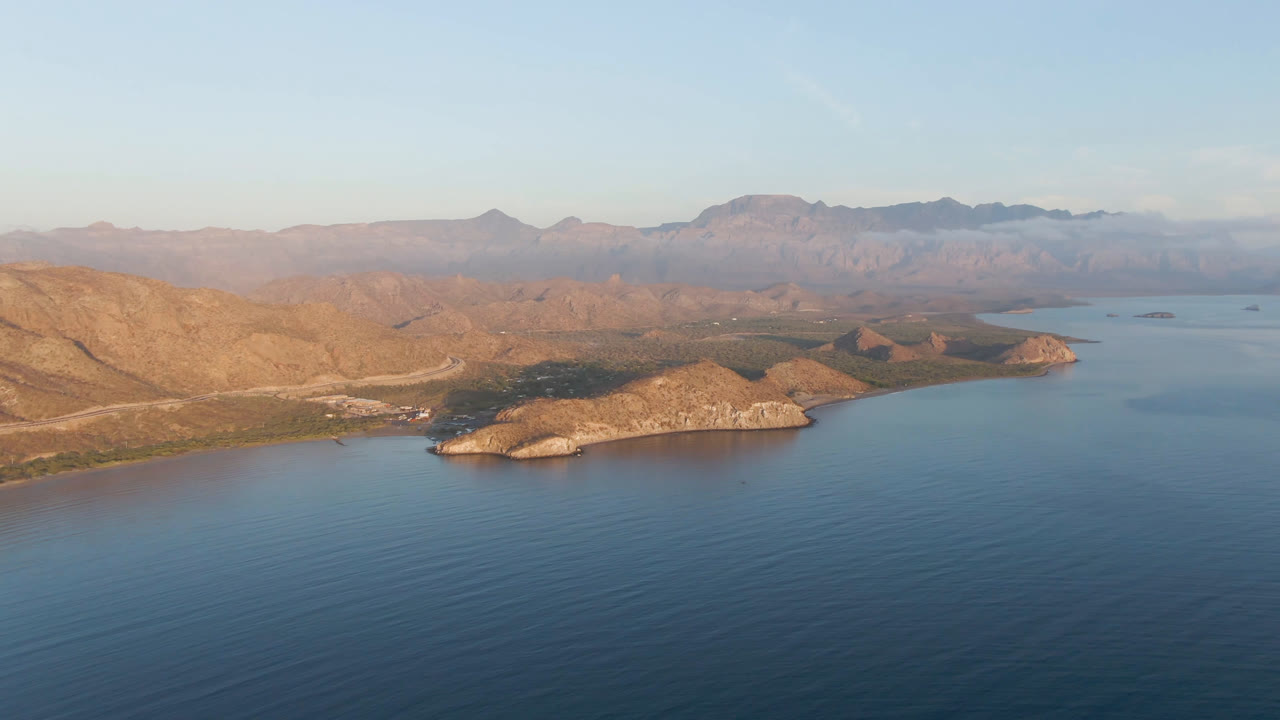 Wide drone-like view of coastal hills, blue water, and layered mountains in haze.
