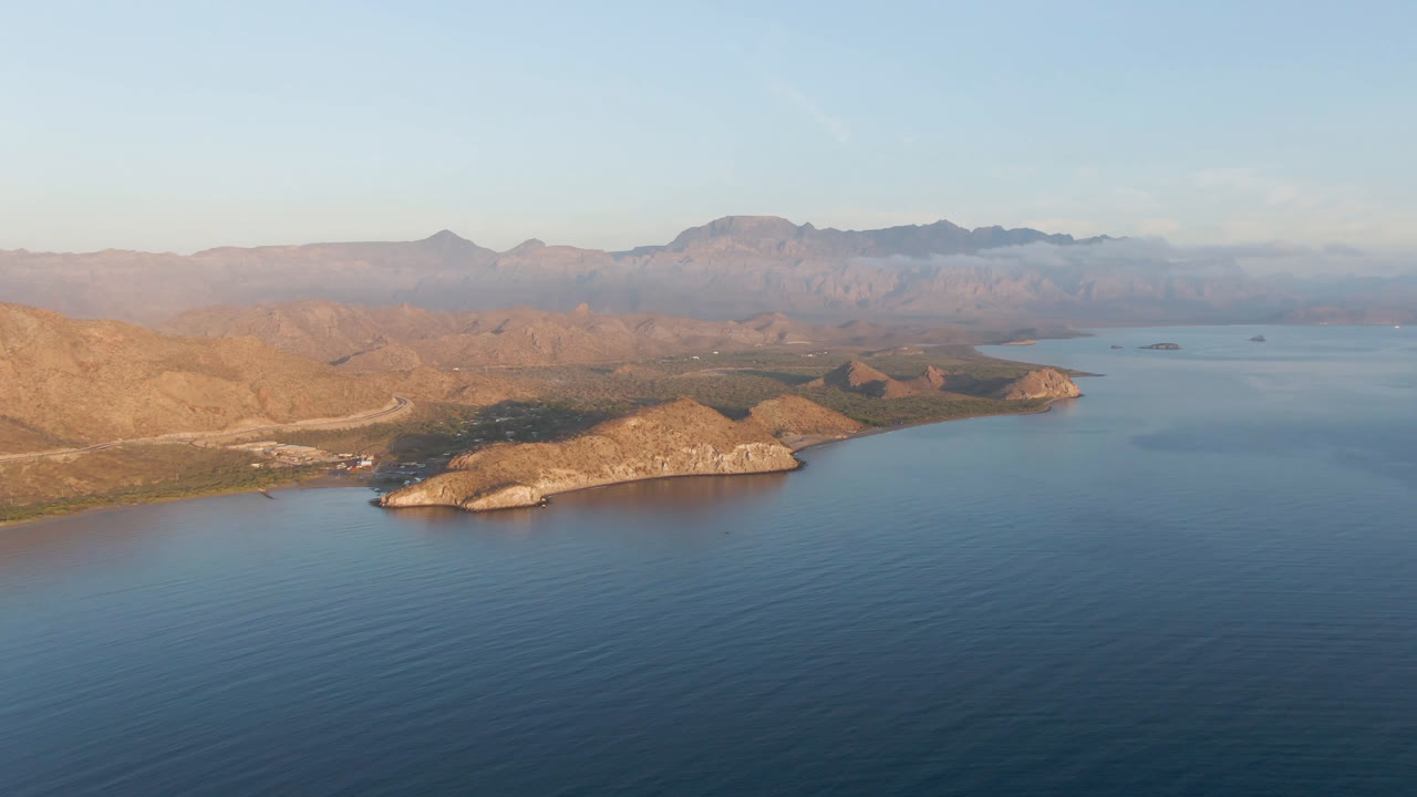 Wide aerial frame of Danzante Bay in Loreto, Baja California Sur, with blue water in front, rocky points along the shore, distant desert mountains, clear sky, and no visible people.