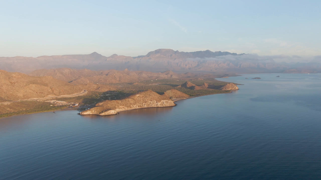 Aerial view over Danzante Bay, Loreto, showing calm blue water, pale rocky shoreline, desert hills, distant mountain ridges, a clear horizon, and no visible people.