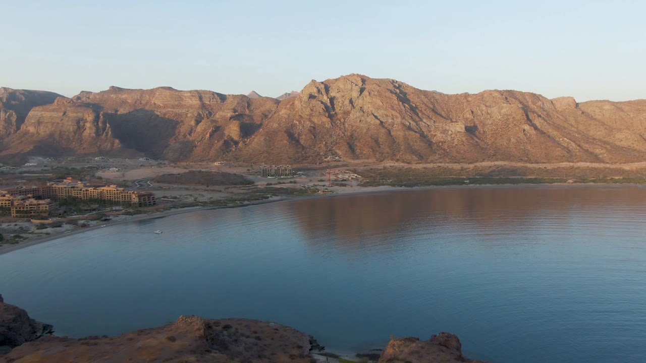 Danzante Bay Loreto aerial frame with calm blue water below, a narrow shoreline across the middle, brown mountain ridges in warm light, and no visible people.