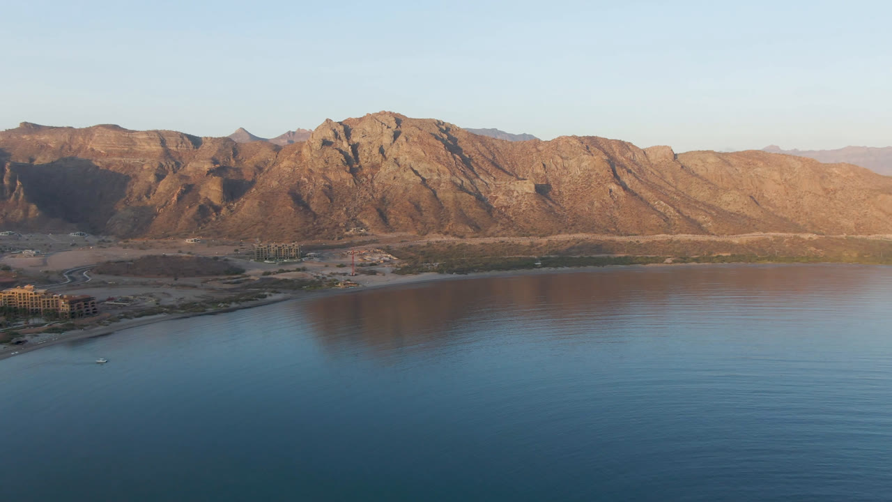 Danzante Bay Loreto seen from above with calm blue water, a small boat near the shoreline, low structures at the left edge, warm-lit mountains, and no visible people.