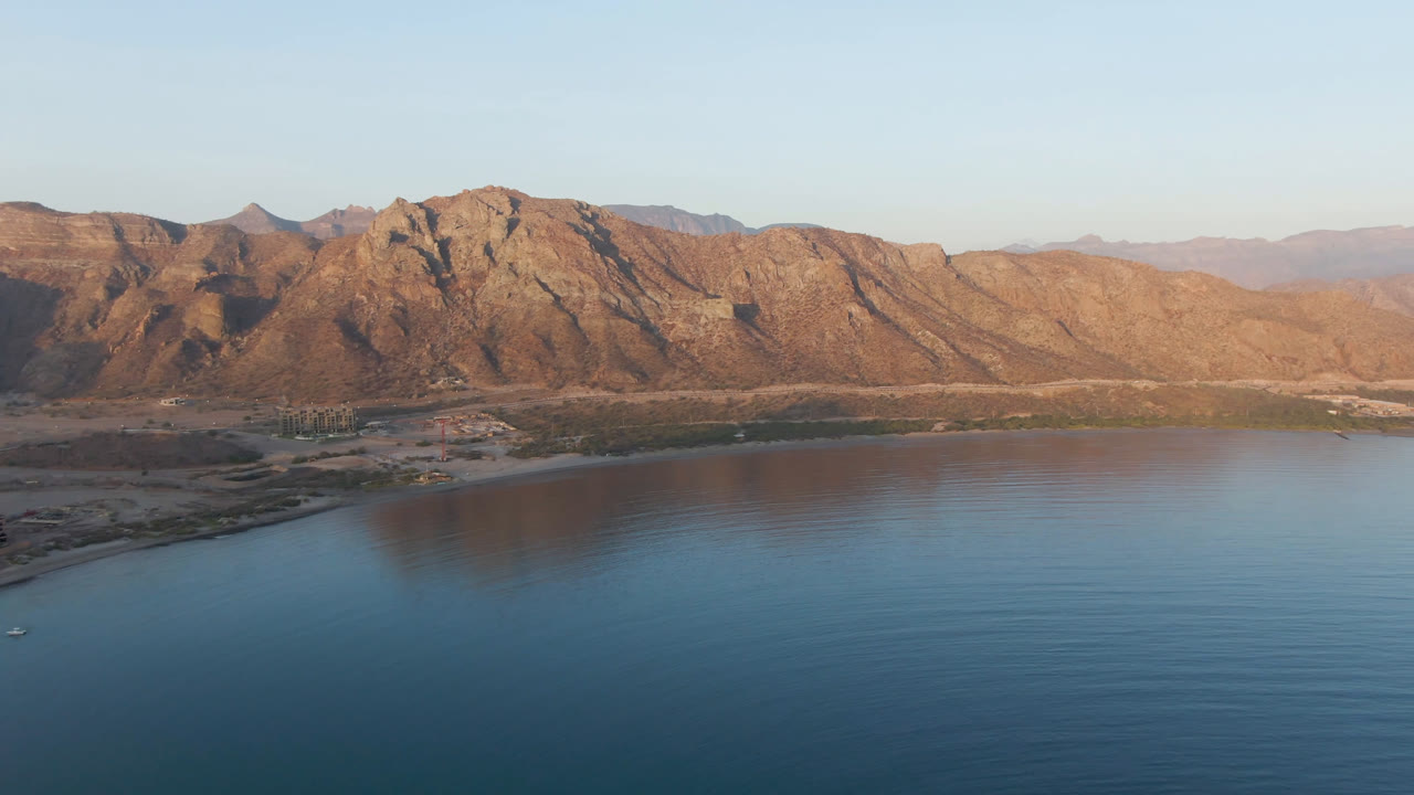 Opening aerial frame over Danzante Bay, Loreto, Baja California Sur, with blue water in the foreground, a thin shoreline, tan mountains in warm light, and no visible people.