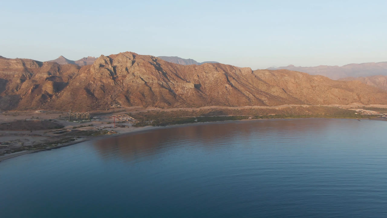 Aerial frame over Danzante Bay showing slightly more shoreline detail below the sunlit mountain ridge, with calm blue water in front and no visible people.