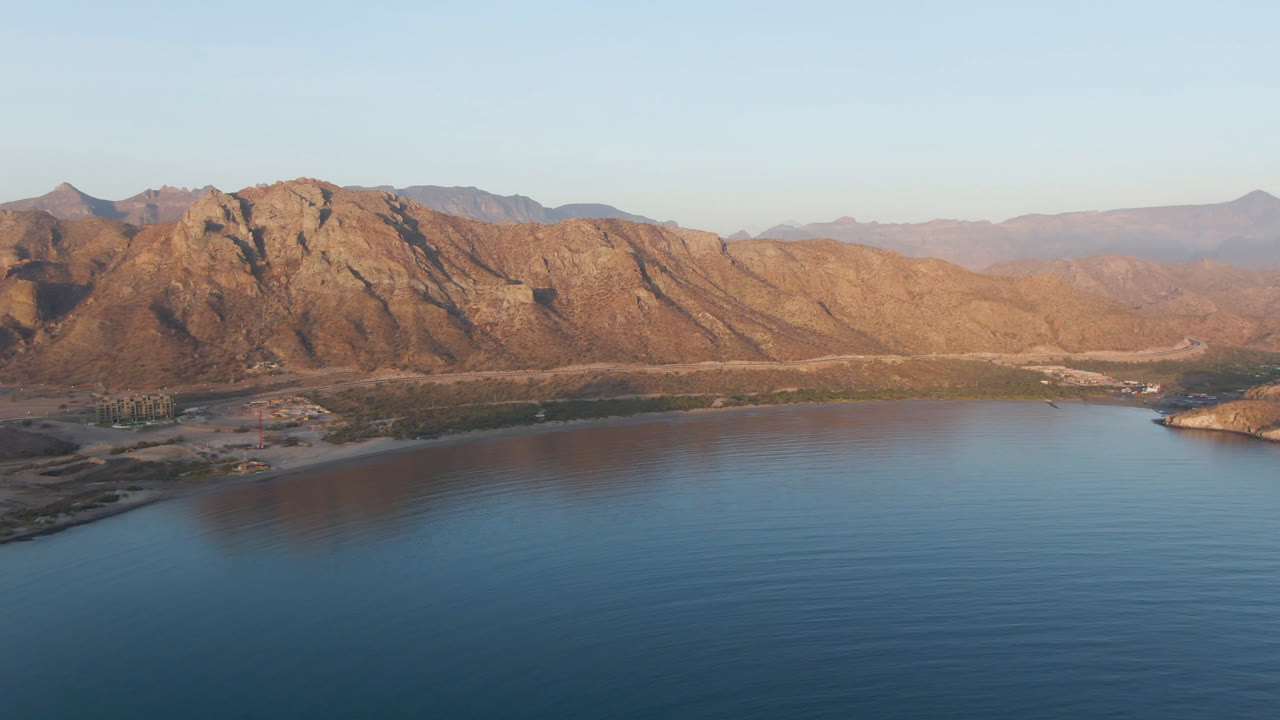 Wide aerial frame over Danzante Bay in Loreto, Baja California Sur, with blue water in the foreground, a low shoreline, sunlit brown mountains, and no visible people.