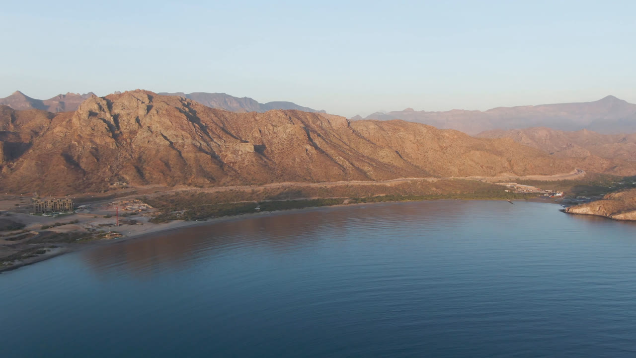 Aerial view of Danzante Bay showing calm blue water, a curving shoreline, rugged brown mountain slopes, pale sky, and no visible people.