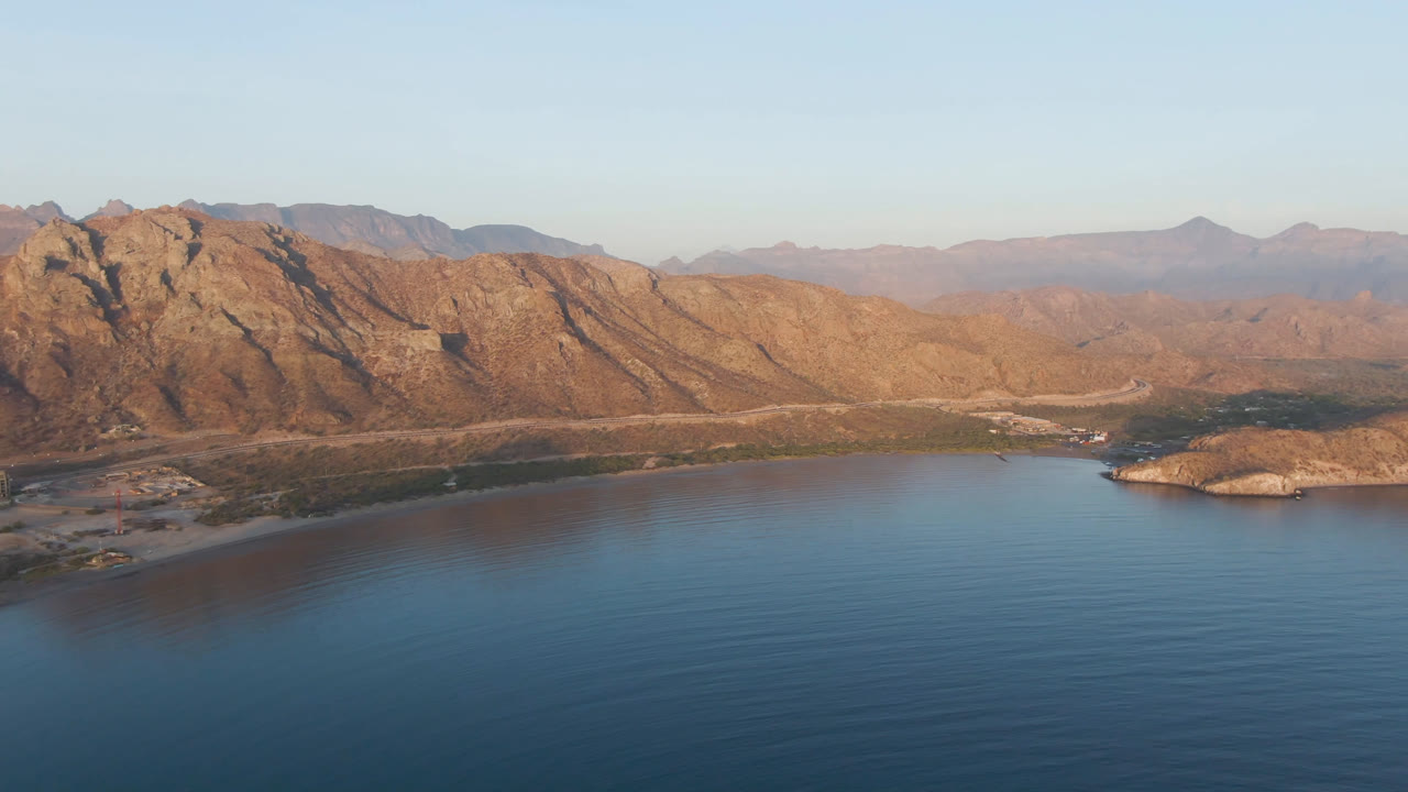 Opening aerial view of Danzante Bay in Loreto, Baja California Sur, with blue bay water, a small rocky island near the shoreline, layered brown mountains, and no visible people.