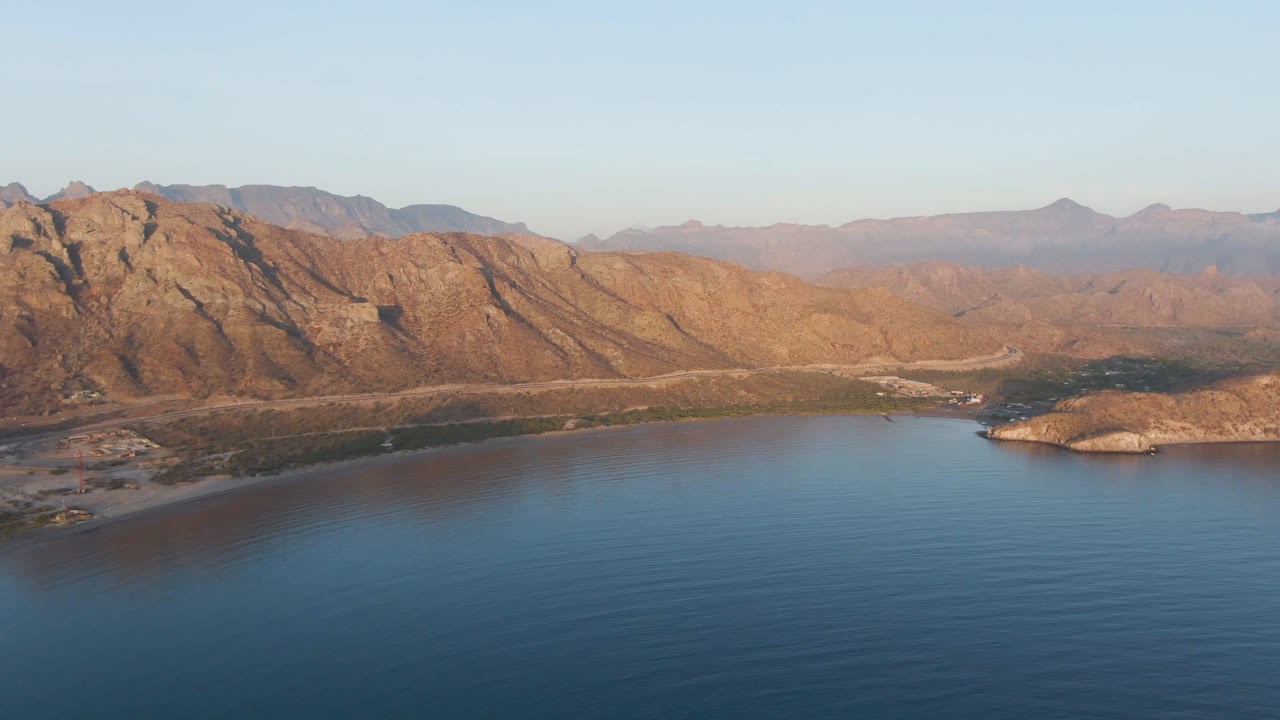 Aerial frame over Danzante Bay showing calm blue water in the foreground, the small rocky island closer to center, sunlit mountain ridges behind it, and no visible people.